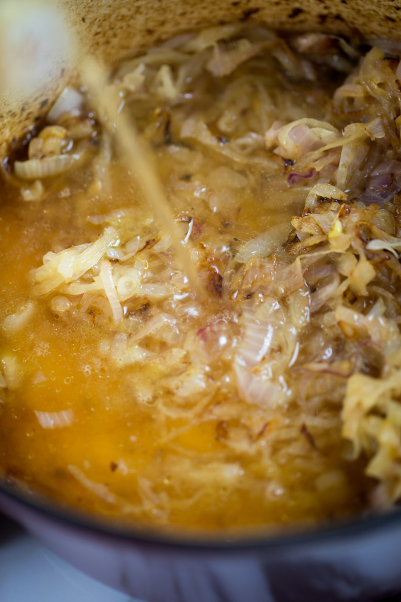 Close-up of onions being caramelized in a pot. The golden brown onions are submerged in a light brown broth, reminiscent of a rich onion soup, with bubbles visible. The pot has a rustic, brown interior. Steam rises from the mixture, suggesting heat and ongoing cooking. A liquid stream is being poured in.
