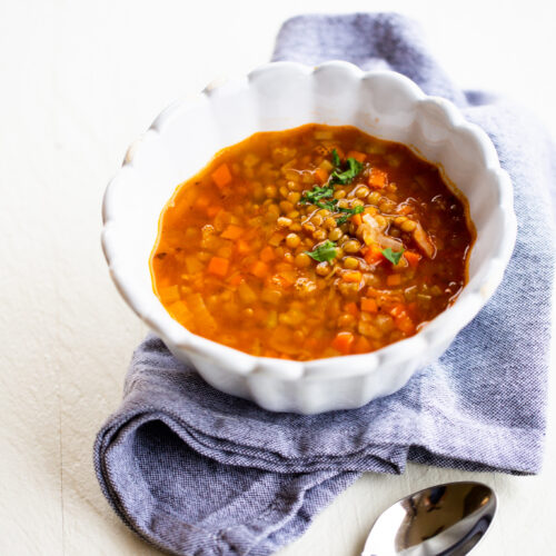 A white scalloped bowl filled with Mexican lentil soup, garnished with herbs, sits on a folded light blue cloth napkin. Visible ingredients include orange carrots and green lentils. A metal spoon with a wooden handle rests nearby on a white surface.