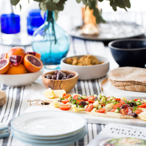 Una mesa luminosa, preparada para una comida, incluye hummus con ensalada, pan plano con verduras, un bol de naranjas sanguinas, aceitunas, pita, falafel, vasos azules, platos blancos y un jarrón de hojas verdes como centro de mesa sobre un mantel a rayas. Un libro de cocina se encuentra en primer plano.