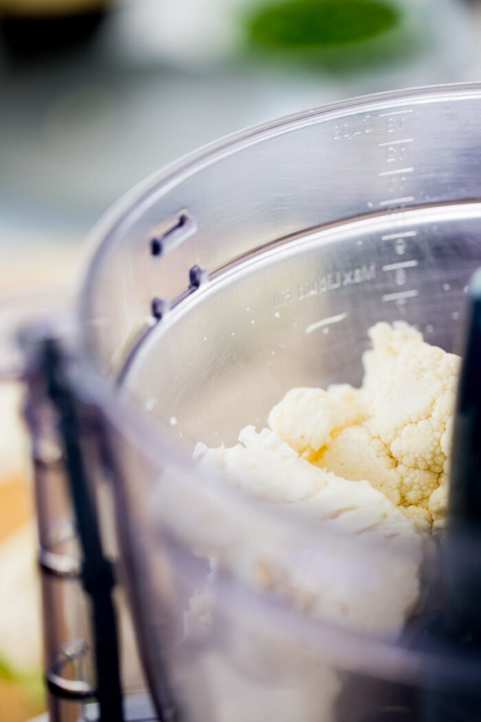 Close-up of cauliflower florets in a transparent food processor bowl, ready to be transformed into Cauliflower Fried Rice. The focus is on the florets and measurement markings, emphasizing fresh food preparation in a home kitchen setting.