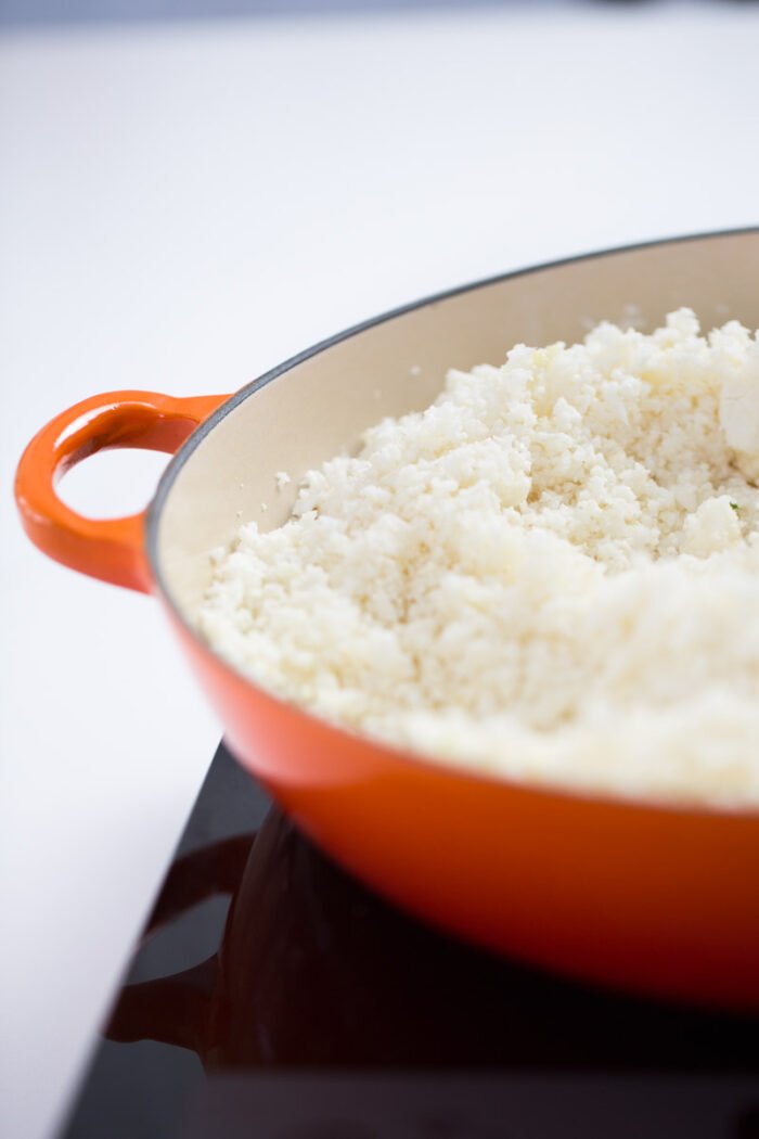 Close-up of a red-orange enameled skillet on a black stovetop, filled with cooked Cauliflower Fried Rice. The skillet&rsquo;s handle is visible on the left, and the blurred background draws focus to the vibrant food and cookware.