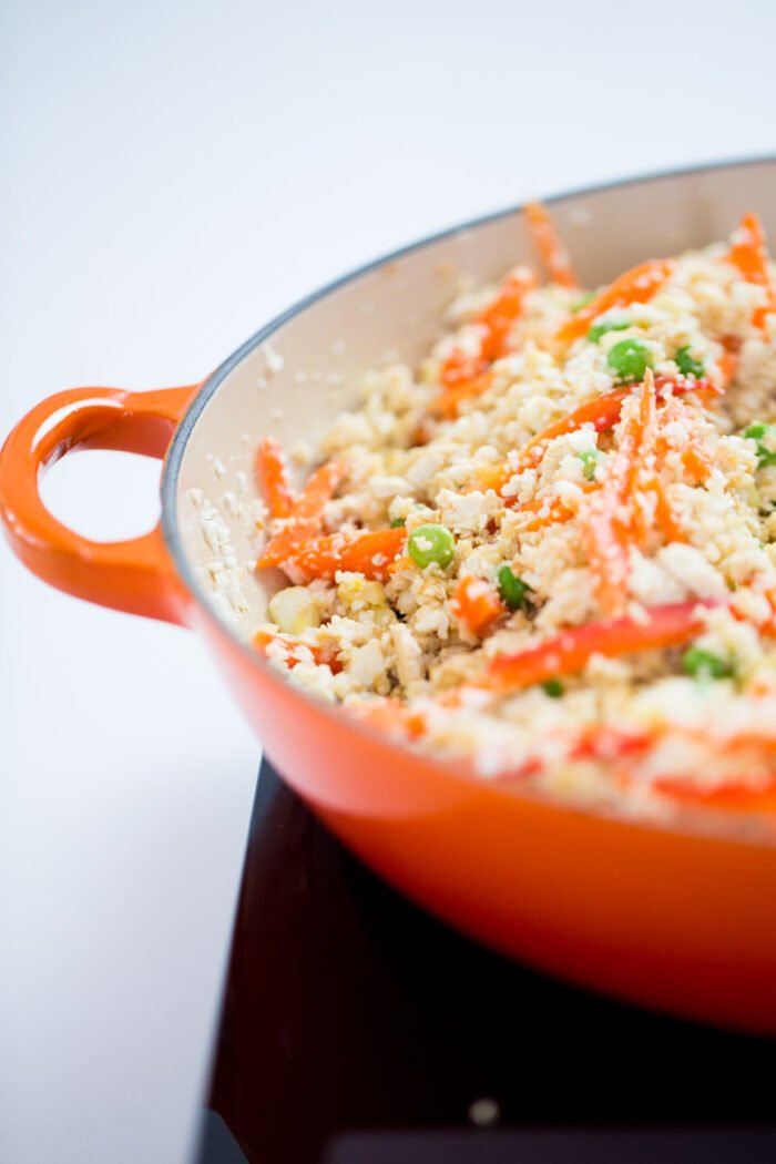 Close-up of an orange-handled skillet filled with colorful cooked vegetables, crumbled white cheese or tofu, and small grains, creating a healthy stir-fry style dish that resembles Cauliflower Fried Rice.
