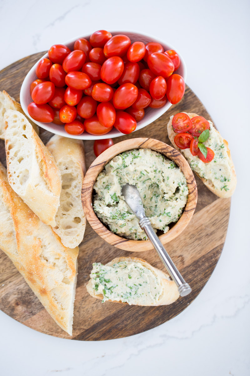 vegan cream cheese with herbs in a wood bowl with cherry tomatoes and sliced baguette on the side.
