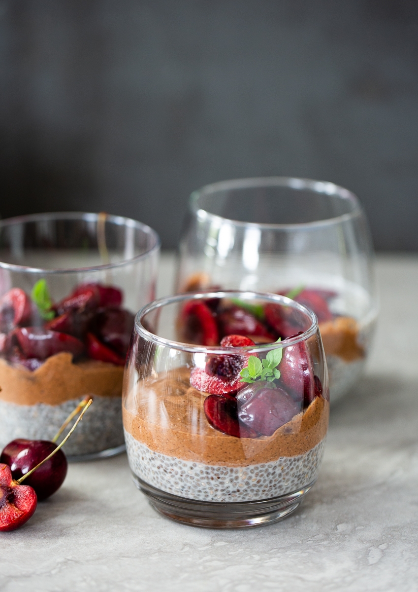 Three clear glasses showcase a beautiful three layer chia pudding with chocolate mousse and cherries, garnished with mint. Two fresh cherries rest beside the glasses, while a soft, blurred background draws focus to the dessert.