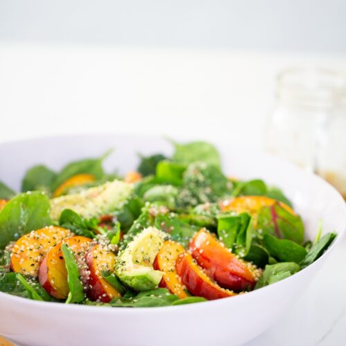 A white bowl brimming with fresh spinach leaves, sliced red and yellow tomatoes, and sprinkled with seeds, likely hemp hearts. A wooden fork peeks into the scene, while a blurred glass jar of vinaigrette sits in the background on a light surface—a refreshing nod to summer peach salad vibes.
