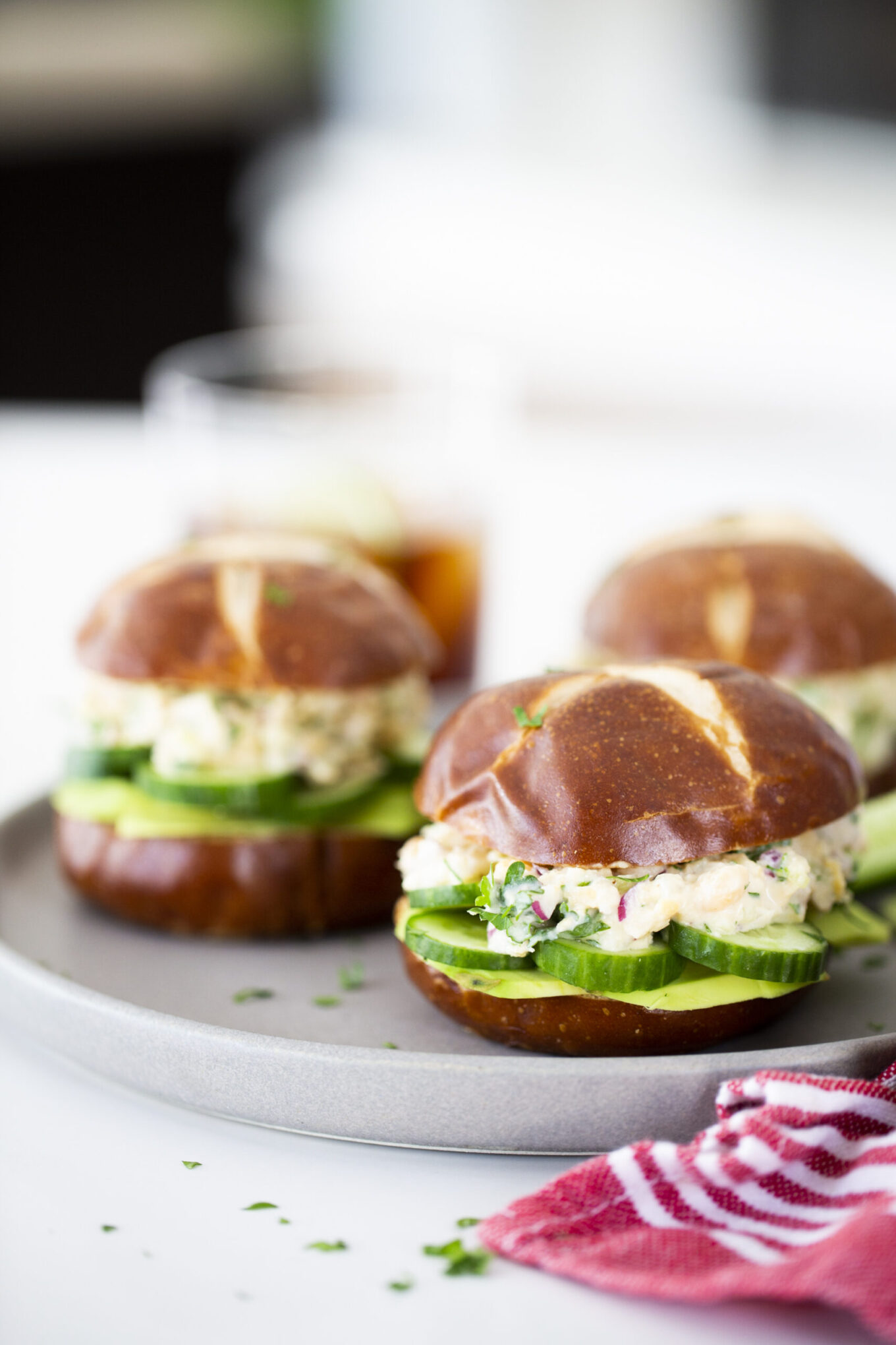 Three pretzel bun sandwiches filled with sliced cucumber and a creamy tuna-less sandwich mixture are arranged on a round gray plate. A red-striped napkin and a beverage in a glass complete the bright, casual setting.