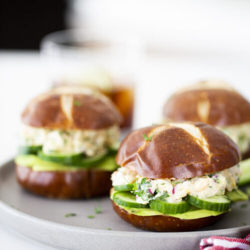 Three pretzel bun sandwiches filled with sliced cucumber and a creamy tuna-less sandwich mixture are arranged on a round gray plate. A red-striped napkin and a beverage in a glass complete the bright, casual setting.
