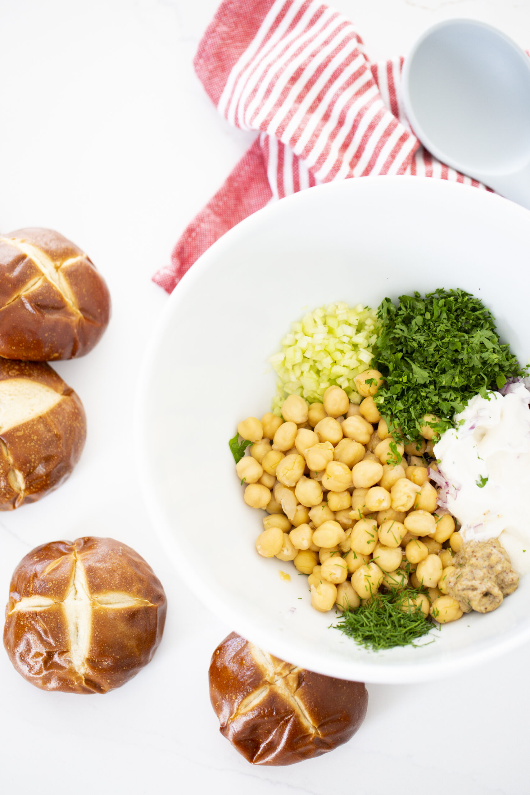 A white bowl contains chickpeas, diced celery, chopped parsley, chopped dill, diced red onion, mayonnaise, and mustard—perfect for a tuna-less sandwich. Next to the bowl are three pretzel sandwich rolls and a red-and-white striped towel on a white surface.