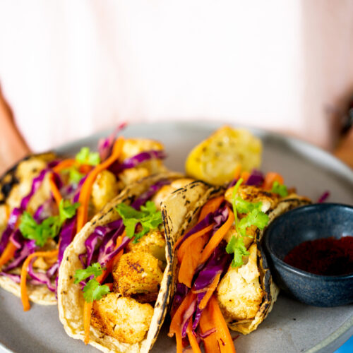 A person holds a plate with three chipotle roasted cauliflower tacos filled with shredded purple cabbage and carrot strips, garnished with fresh cilantro. A lemon wedge and a small black bowl with a red spice accompany the vibrant dish. The person wears a light shirt and blue jeans.