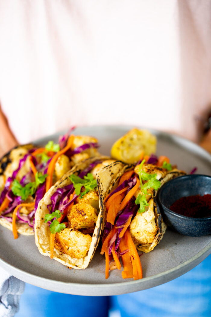 A person holds a plate with three chipotle roasted cauliflower tacos filled with shredded purple cabbage and carrot strips, garnished with fresh cilantro. A lemon wedge and a small black bowl with a red spice accompany the vibrant dish. The person wears a light shirt and blue jeans.