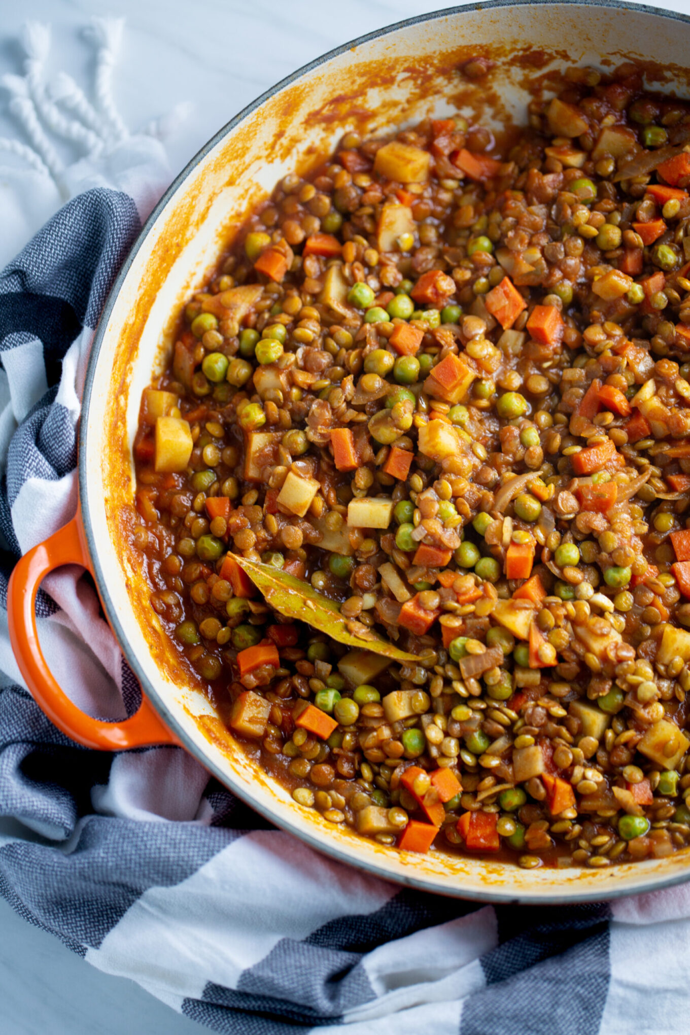 A close-up of a white pot with an orange handle filled with hearty lentil picadillo stew containing peas, carrots, potatoes, and a bay leaf. The pot rests on a black, white, and gray striped cloth on a white surface.