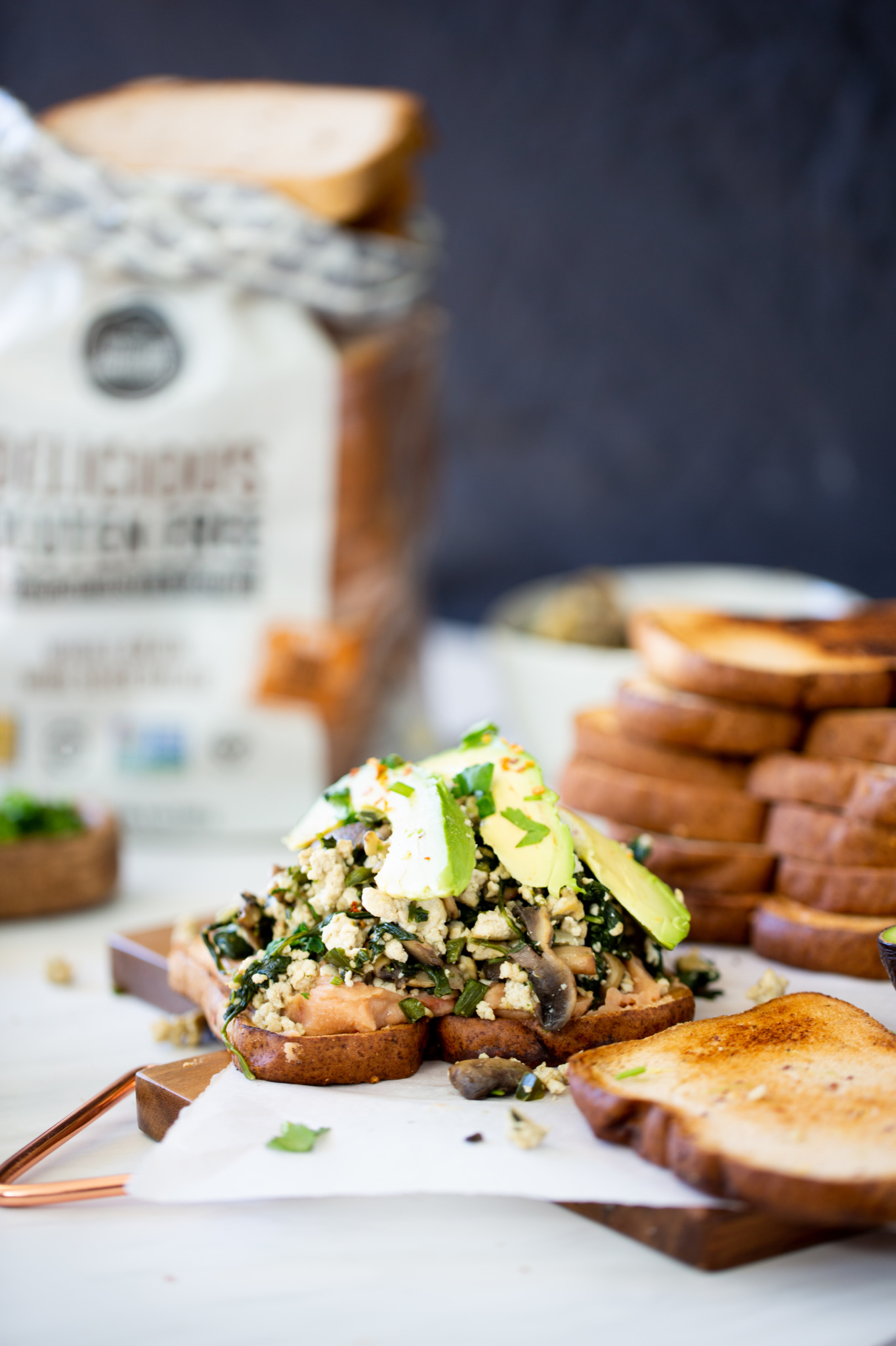 Dos rebanadas de pan tostado se cubren con verduras salteadas, champiñones, queso feta desmenuzado y rodajas de aguacate para un sándwich repleto de proteínas. Más rebanadas de pan y una barra de pan en una bolsa se sientan en el fondo en una escena brillante tabla de cortar de madera.