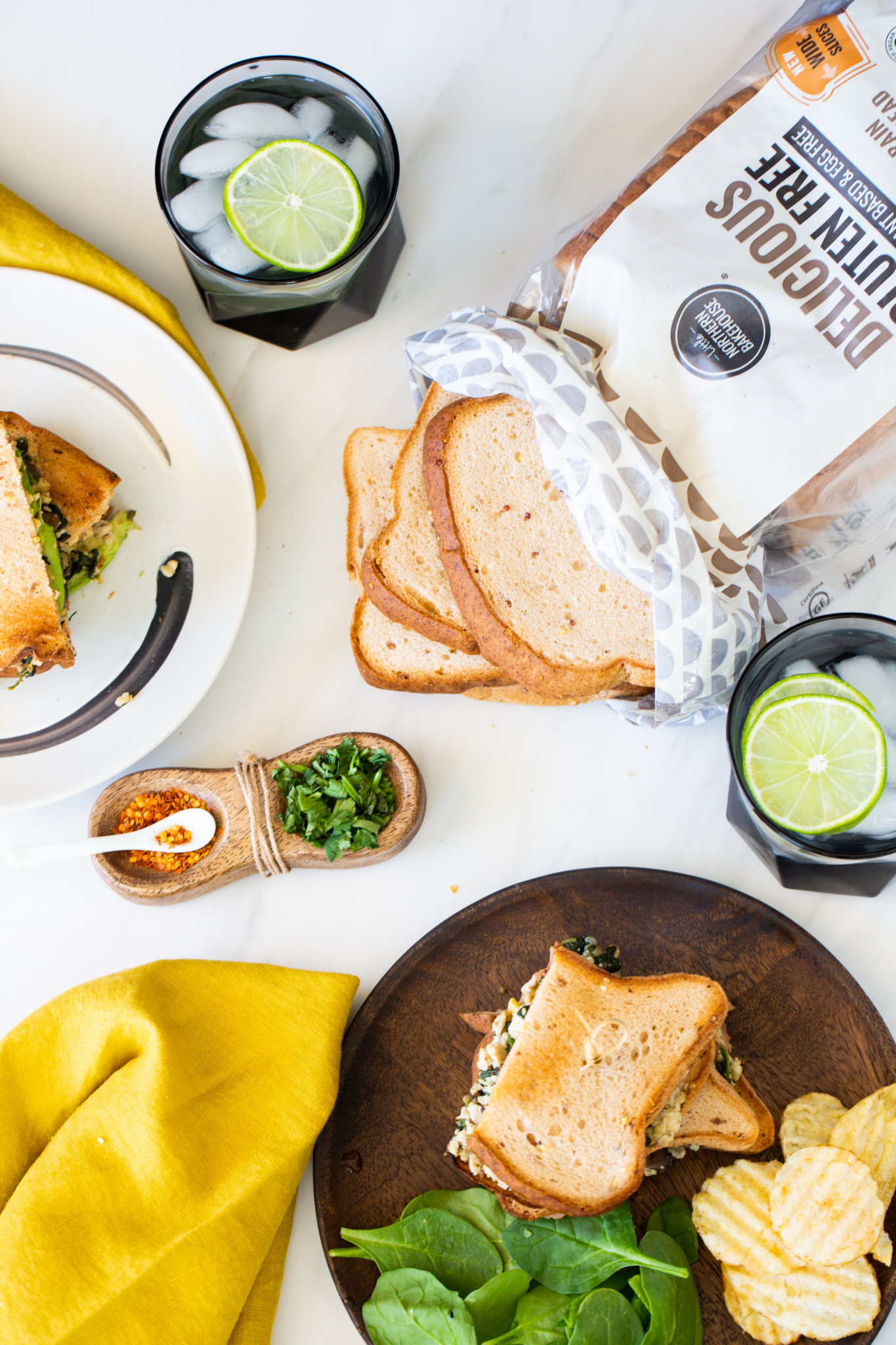 Una vista de arriba abajo de una comida de sándwich repleta de proteínas con pan sin gluten, dos sándwiches en platos, espinacas frescas, patatas fritas, dos vasos con hielo y lima, una servilleta amarilla y pequeños cuencos con hierbas picadas y condimentos sobre una superficie blanca.