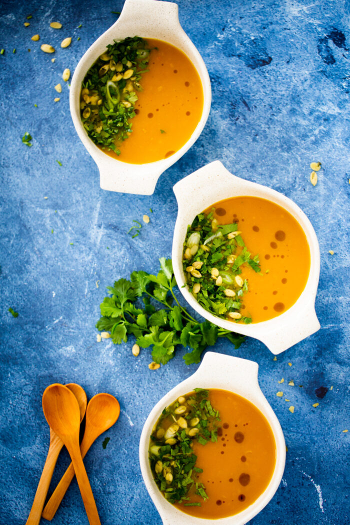 Three white bowls of carrot ginger detox soup sit on a blue textured surface. The vibrant soup is garnished with chopped herbs and seeds, while fresh cilantro adds a fresh touch between the bowls and wooden spoons placed nearby.