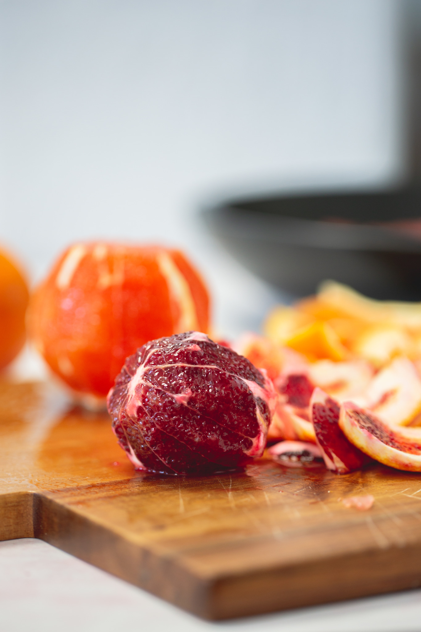 A peeled blood orange sits on a wooden cutting board in the foreground, its deep red flesh exposed—perfect for a citrus salad. Behind it are orange peels and another partially peeled orange, with a softly blurred background drawing focus to the fruit.