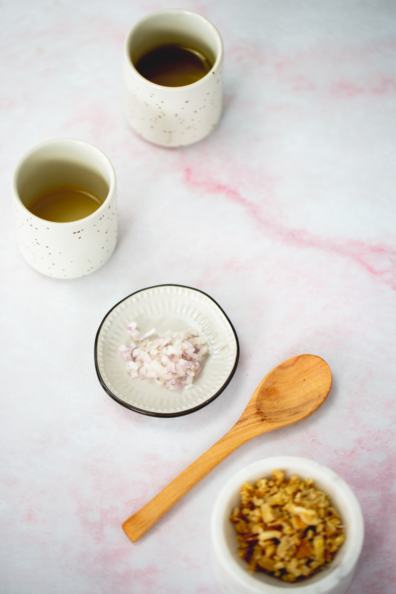 Two speckled white cups with tea, a small plate of finely chopped shallots, a wooden spoon, and a bowl of chopped walnuts are arranged on a light pink marbled surface, evoking the fresh vibe of a citrus salad. The items are neatly placed and softly lit.