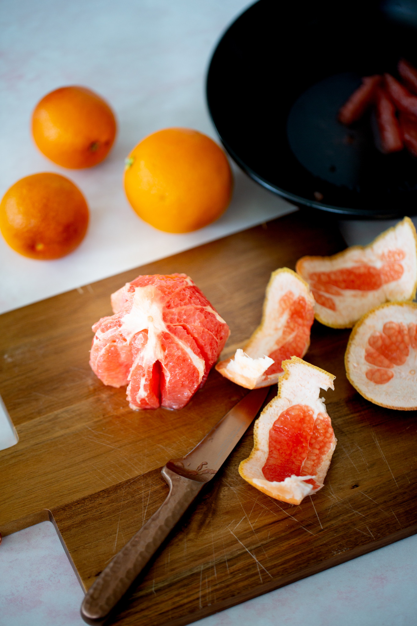 A peeled grapefruit sits on a wooden cutting board next to a knife and grapefruit peels. Three unpeeled oranges are nearby, hinting at citrus salad prep, while a black bowl with grapefruit segments is in the background.