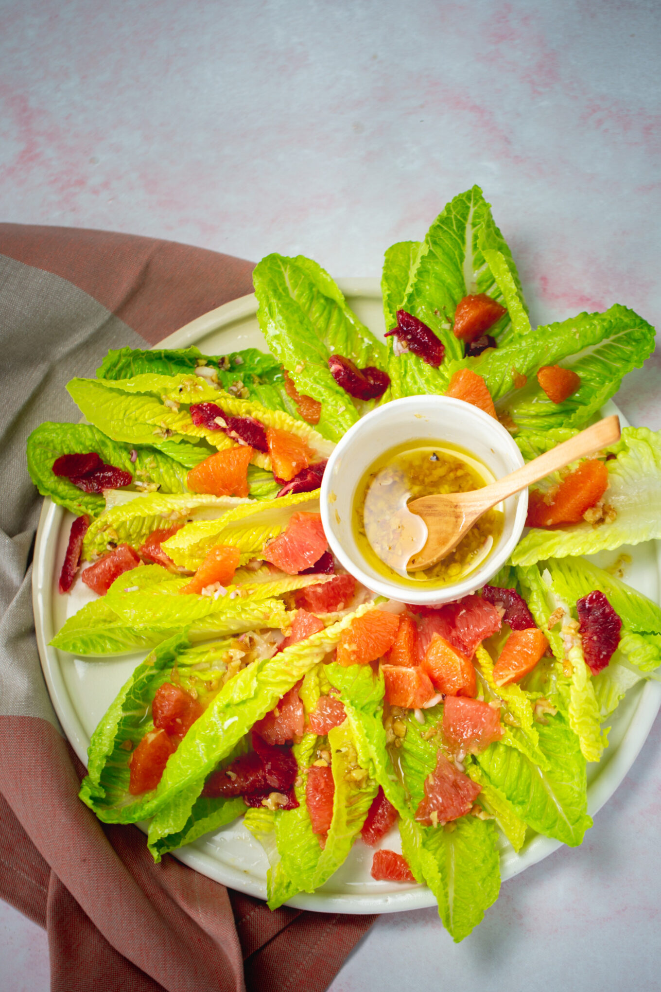 A plate of romaine lettuce leaves arranged in a circle forms a vibrant citrus salad, topped with orange and red citrus segments. In the center is a small bowl of vinaigrette with a wooden spoon, set on soft pink and brown cloth against a light pink background.