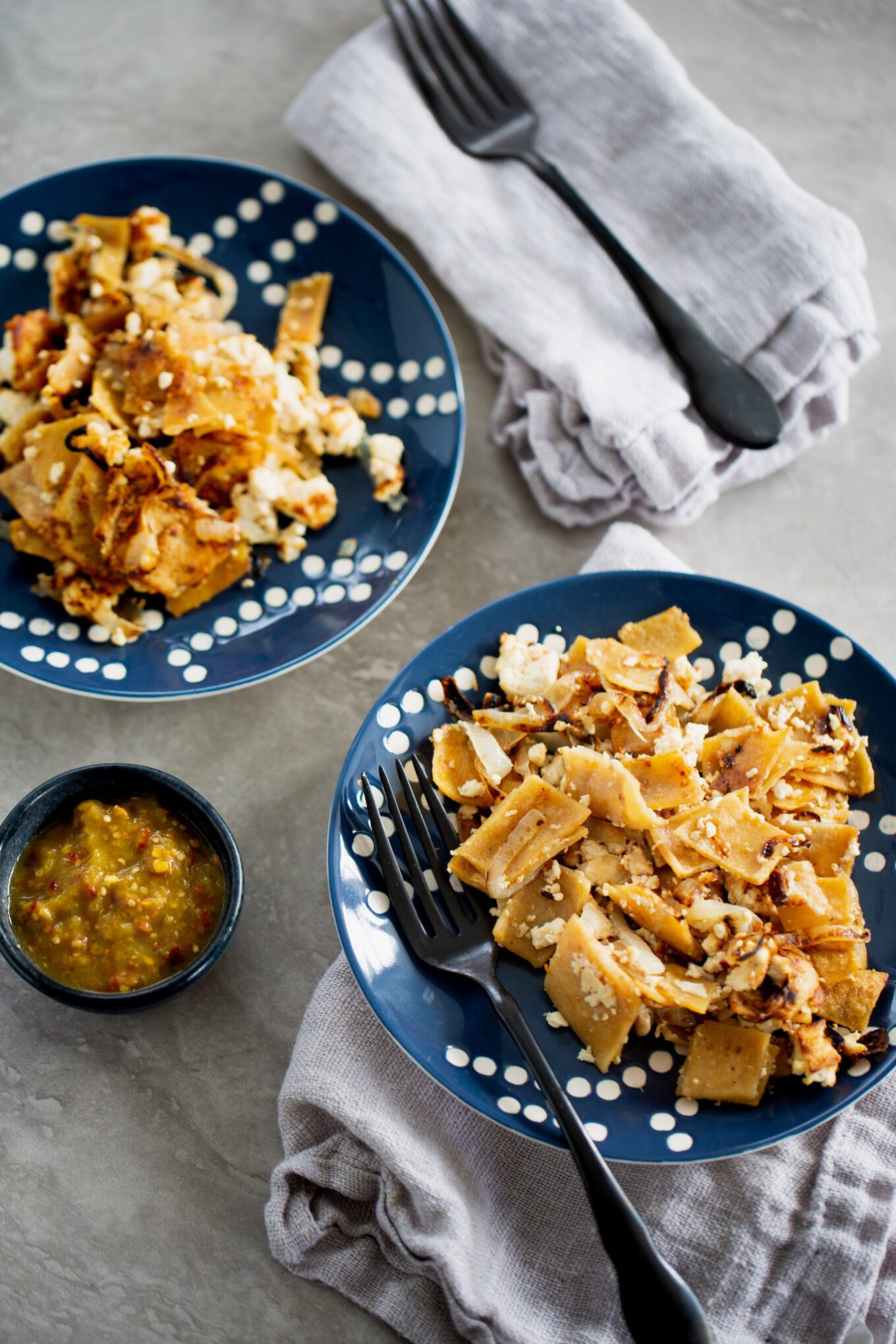 Two blue plates with white dots hold servings of chilaquiles—crispy tortilla pieces mixed with cheese and sauce, similar to vegan migas. Black forks rest on the plates. Nearby, light gray napkins and a small bowl of green salsa sit on a light surface.