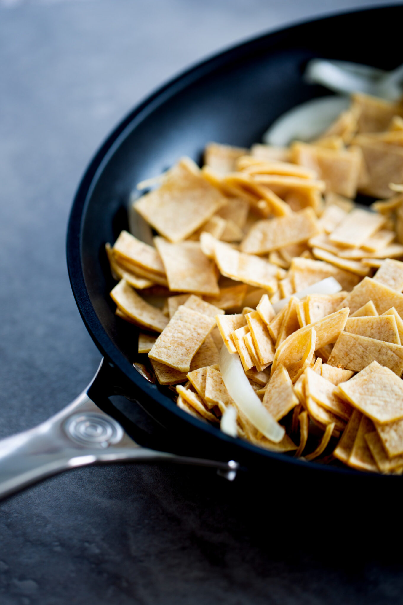 A close-up of a black frying pan with a silver handle, filled with uncooked tortilla strips and sliced onions—perfect for preparing vegan migas—rests on a dark gray surface. The light brown tortilla strips are cut into small squares and rectangles.