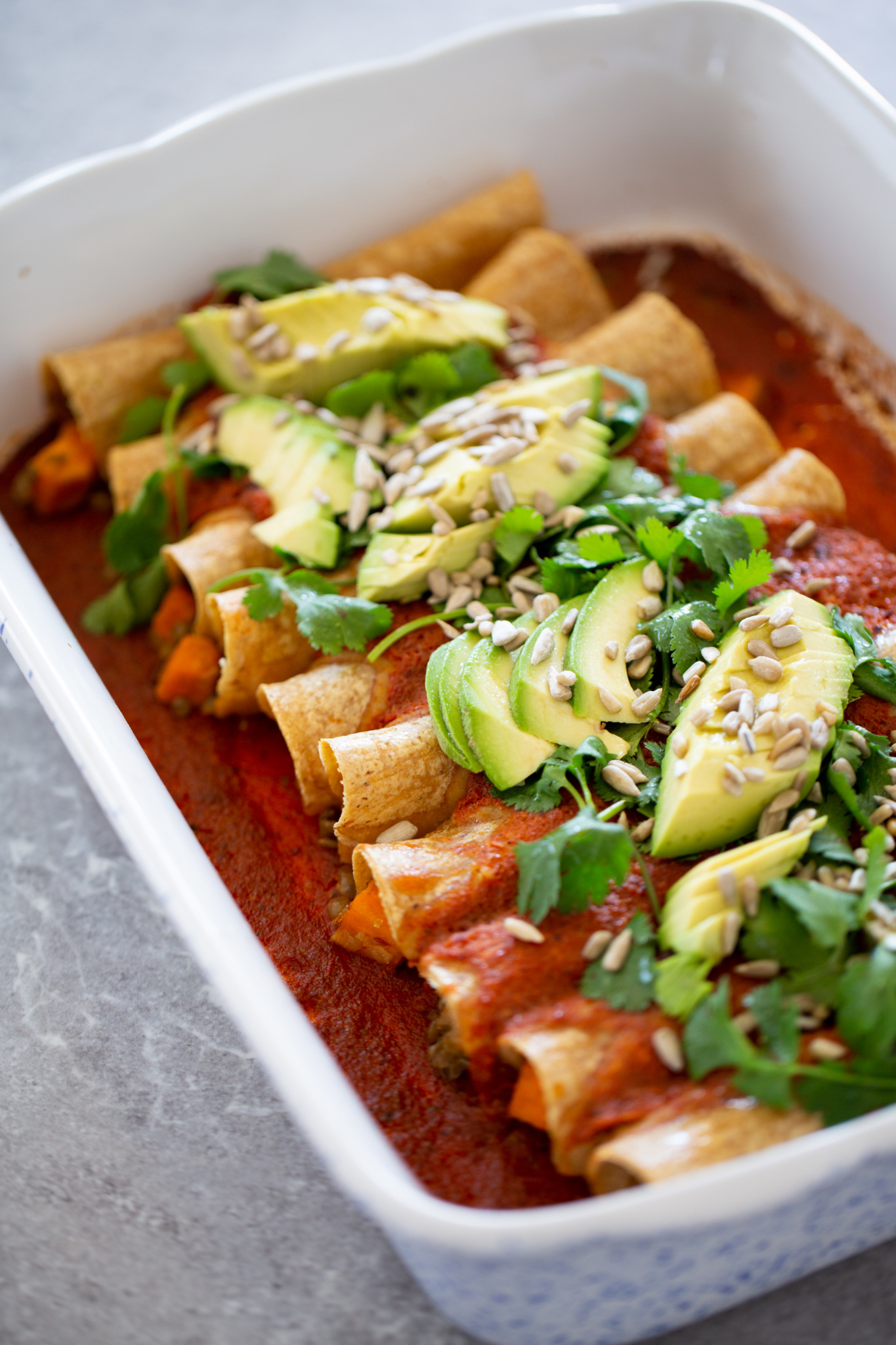 A plate of vegan enchiladas topped with sliced ​​avocado, cilantro and seeds served in a ceramic baking dish.