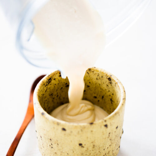 A creamy, beige tofu crema is being poured from a blender into a speckled ceramic cup. A wooden spoon rests beside the cup on a white surface. The liquid forms gentle swirls as it fills the cup in this bright, minimalistic scene.