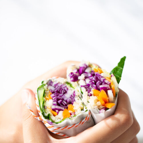 Two hands hold a vibrant collard greens burrito cut in half, revealing layers of purple cabbage, orange bell pepper, and other fresh vegetables wrapped in white paper with a red string, set against a bright, softly-lit background.