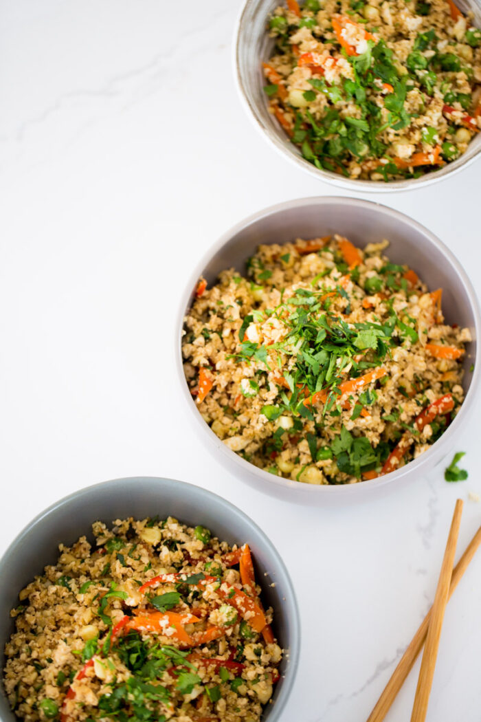 Three bowls of colorful quinoa salad, featuring chopped veggies like red bell pepper and green herbs, are arranged on a white surface. Reminiscent of Cauliflower Fried Rice, the salad is garnished with cilantro, with wooden chopsticks beside one bowl.