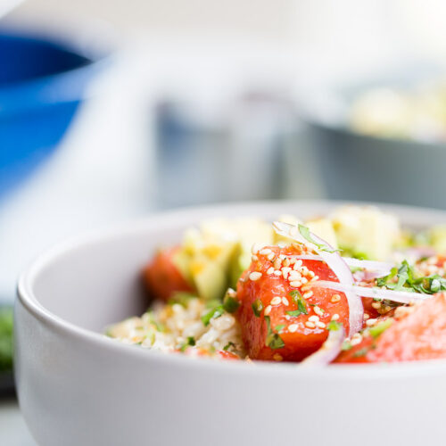 Close-up of a plant-based bowl featuring a fresh salad with cherry tomatoes, diced avocado, sliced onions, and sprinkled sesame seeds. The bowl sits on a light surface with a blurred blue bowl in the background, creating a bright and fresh ambiance.