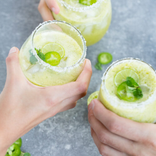 Three hands holding glasses filled with alcohol-free pineapple-mint margaritas, garnished with lime slices and mint. The glasses have salted rims and are brimming with ice. Sliced jalapeños and lime add a vibrant touch to the gray surface below.