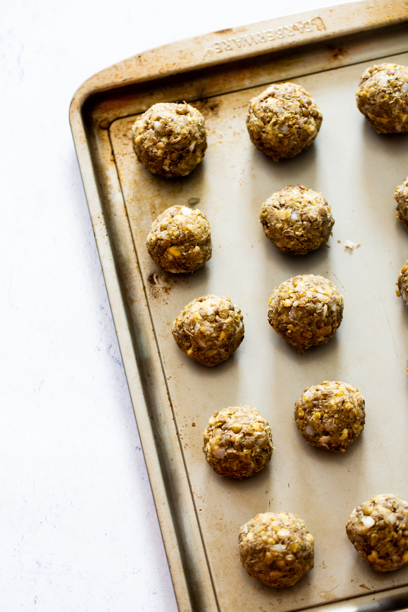 A metal baking sheet holds evenly spaced, raw falafel balls ready to be baked for a vegan meatball sub. The falafel are round, golden-brown, and textured with visible bits of herbs and legumes. The pan sits on a light, white surface.