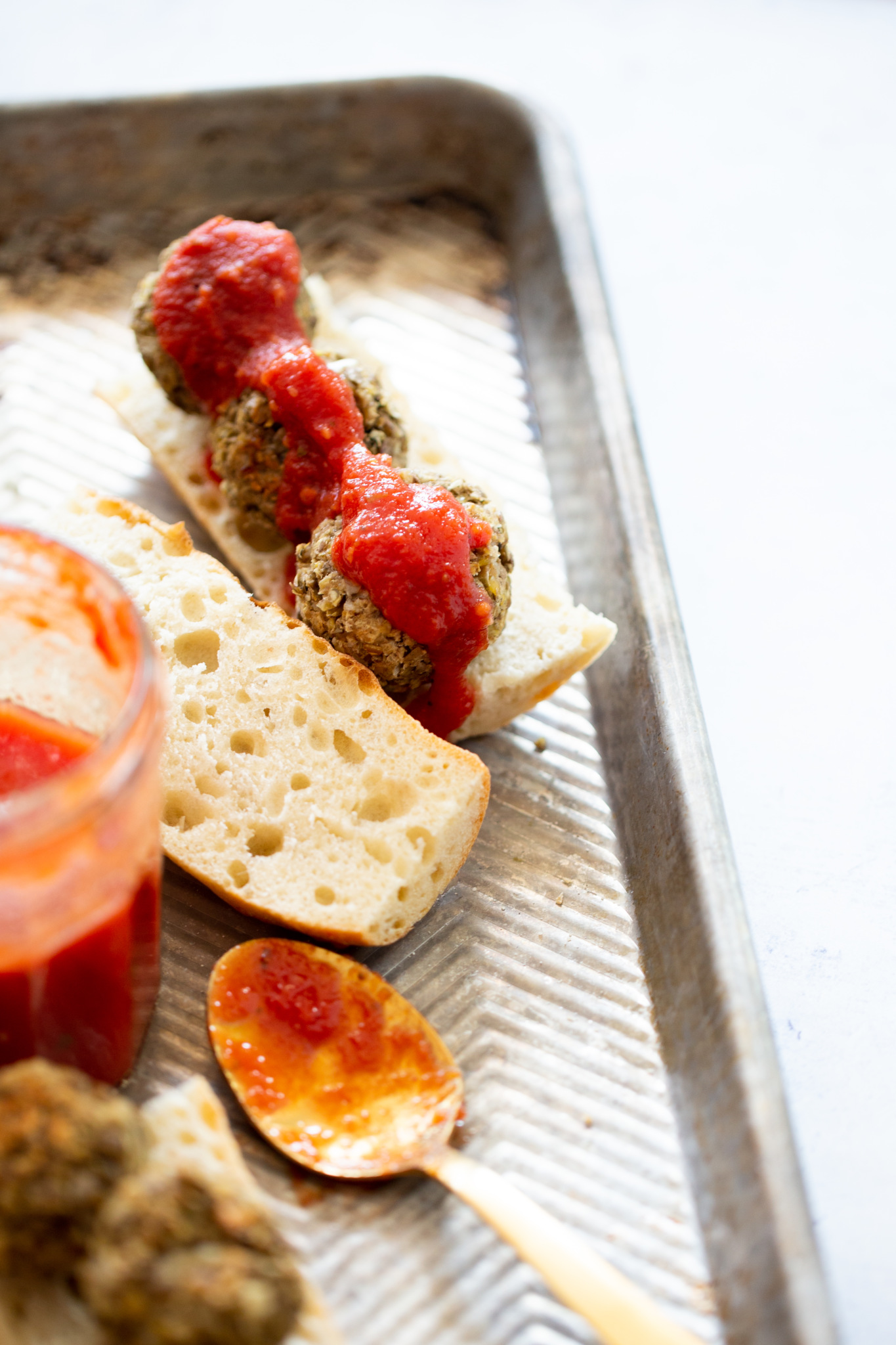 A metal baking tray holds a split piece of bread topped with three vegan meatballs and red tomato sauce. Nearby are a glass jar of sauce, a wooden spoon with sauce, and another vegan meatball, all on a bright, white background.