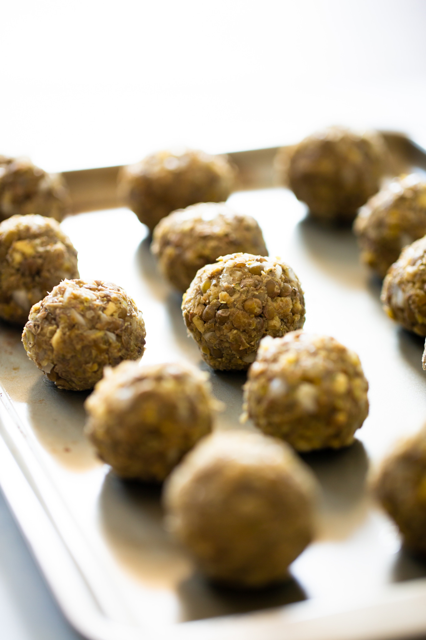 A metal baking tray holds several round, textured energy bites made of oats and seeds, arranged in rows. Their look is reminiscent of vegan meatball sub fillings, with visible oats and ingredients. The softly blurred background highlights the treats.