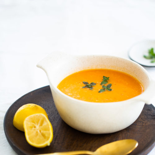 A white ceramic bowl filled with bell pepper and potato soup, garnished with green herbs, sits on a dark wooden plate. Beside the bowl are a halved lemon and a gold spoon. The bowl and plate rest on a white tablecloth, with a small dish of green herbs in the background.