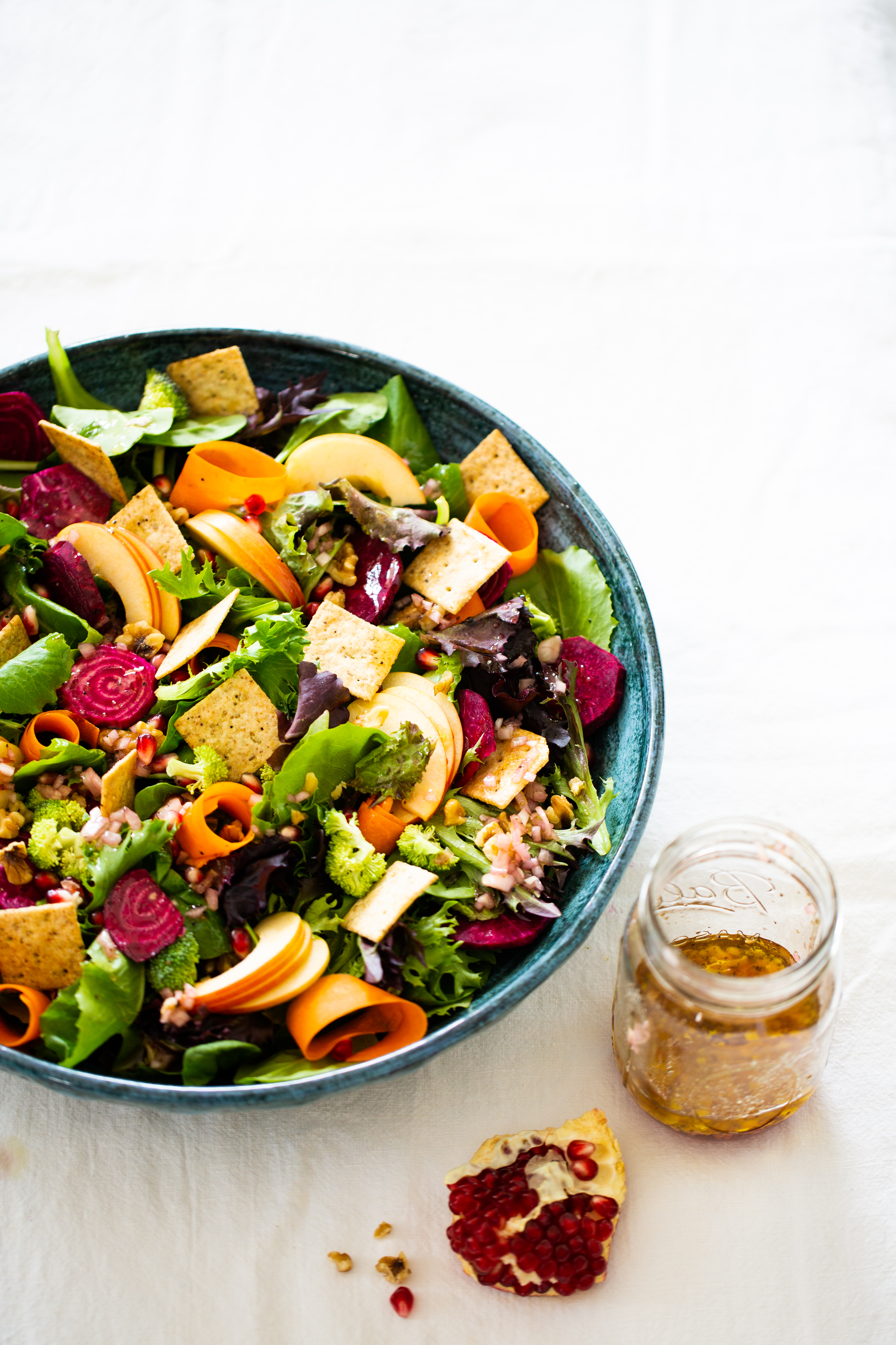 Una vibrante ensalada otoñal en un bol azul con hojas verdes, rodajas de zanahoria, rábanos, pepinos, chips de pita y granos de granada. La ensalada está colocada sobre un mantel blanco con un tarro de cristal con el aliño y granos de granada esparcidos.