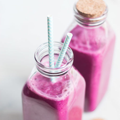 Two glass bottles filled with a vibrant pink beet smoothie are topped with matching cork lids (one removed) and two chevron-patterned blue straws. The bottles sit on a light surface, emphasizing the rich, creamy texture and inviting color of the smoothie.