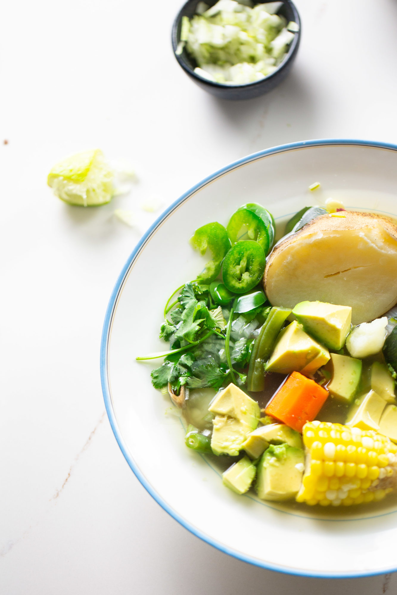 A bowl of soup containing assorted fresh vegetables, including avocado cubes, sliced jalapeños, corn on the cob, and a large piece of fish. Garnished with cilantro. A small bowl with a green garnish reminiscent of vegan Mexican flavors and a squeezed lime wedge are in the background on a white table.