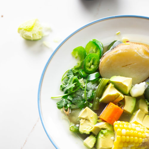 A bowl of soup containing assorted fresh vegetables, including avocado cubes, sliced jalapeños, corn on the cob, and a large piece of fish. Garnished with cilantro. A small bowl with a green garnish reminiscent of vegan Mexican flavors and a squeezed lime wedge are in the background on a white table.