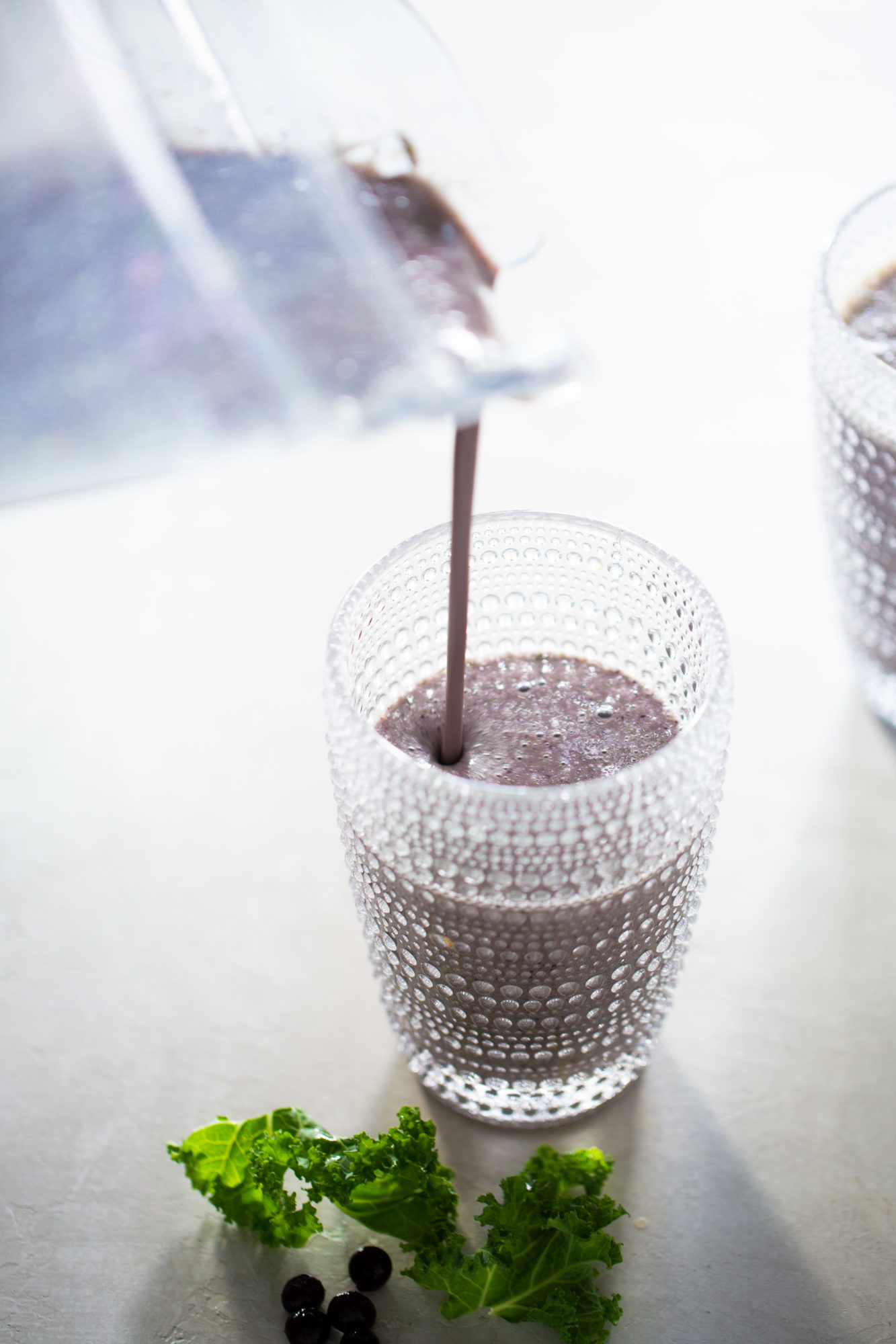A creamy purple smoothie is being poured from a blender into a textured glass. Another identical glass, partially filled with the same favorite blueberry banana smoothie, is visible in the background. Fresh green kale leaves and a few dark blueberries are placed on a light, textured surface next to the glass.