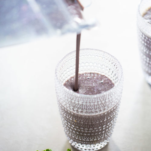 A creamy purple smoothie is being poured from a blender into a textured glass. Another identical glass, partially filled with the same favorite blueberry banana smoothie, is visible in the background. Fresh green kale leaves and a few dark blueberries are placed on a light, textured surface next to the glass.