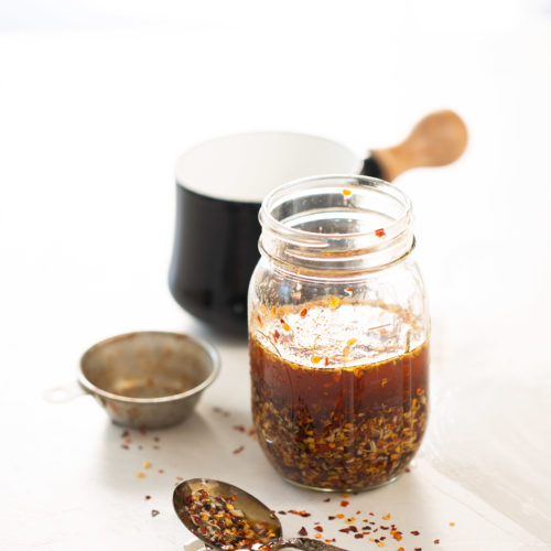 A glass jar filled with a reddish-brown chili crunch sauce and flakes sits on a white surface. Beside it, a metal spoon with the crunchy mixture and a small strainer. In the blurred background, a black mug with a wooden handle is visible. The scene is brightly lit, suggesting a kitchen setting.