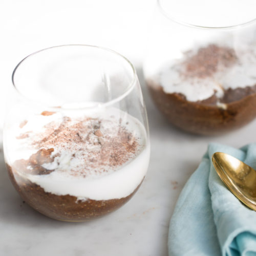 Two clear glasses filled with mocha chia pudding topped with white cream and cocoa powder sit on a light marble surface. A folded light blue cloth lies beside them with a gold spoon resting on top. The background is softly blurred, highlighting the elegance of this dessert arrangement.