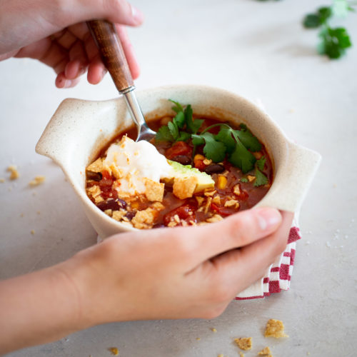 Hands holding a ceramic bowl of tomato soup garnished with fresh cilantro, crumbled crackers, and a dollop of sour cream evoke the comforting warmth of a three bean chili. A wooden-handled spoon rests inside the bowl on a red and white checkered cloth, with spilled crumbs and a cilantro sprig scattered whimsically.