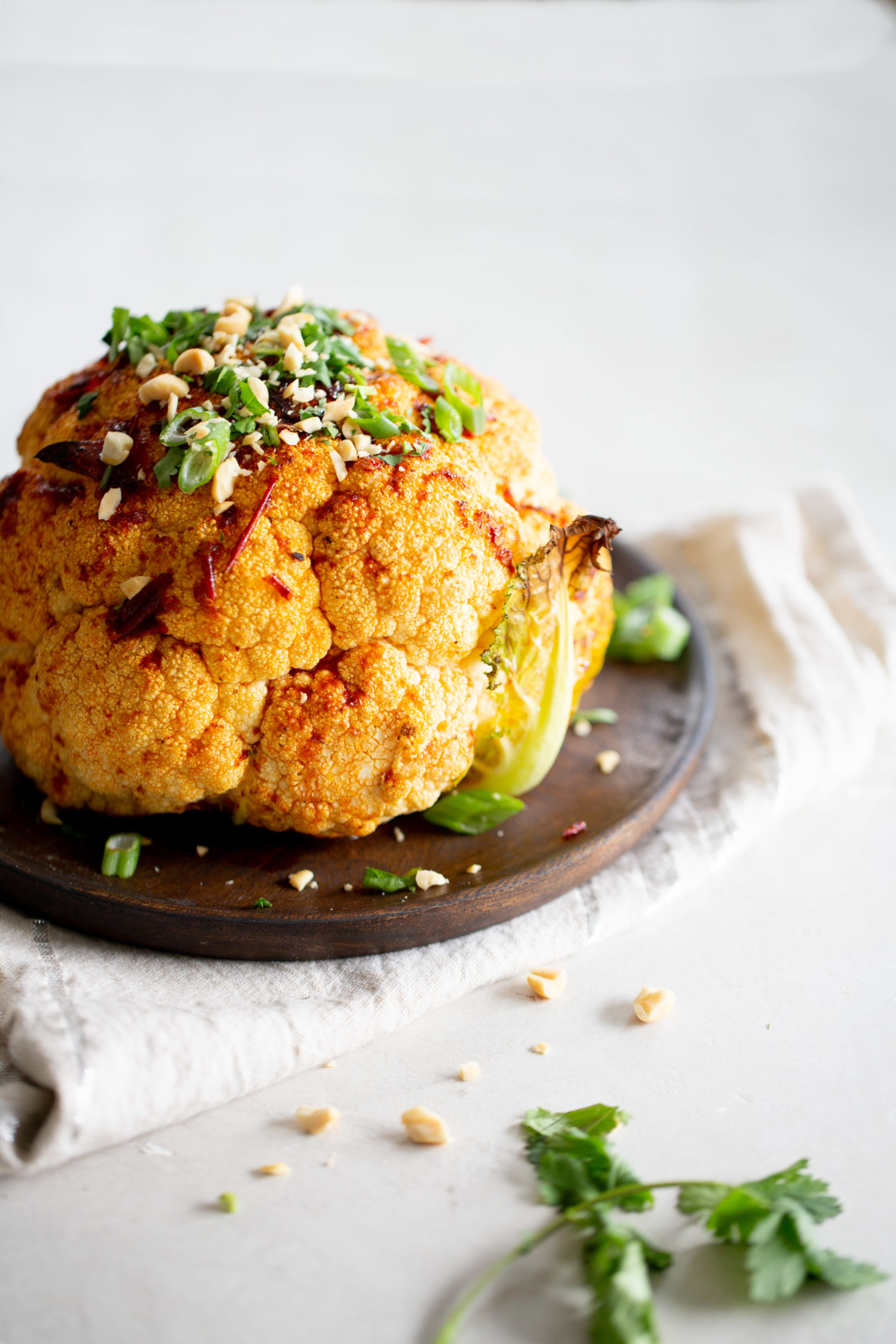 A roasted cauliflower sits on a rustic wooden plate, seasoned with spices and topped with green herbs, chopped nuts, and slices of green onions. A light-colored cloth is underneath the plate, with scattered herbs and nuts surrounding the roasted cauliflower on the white surface.