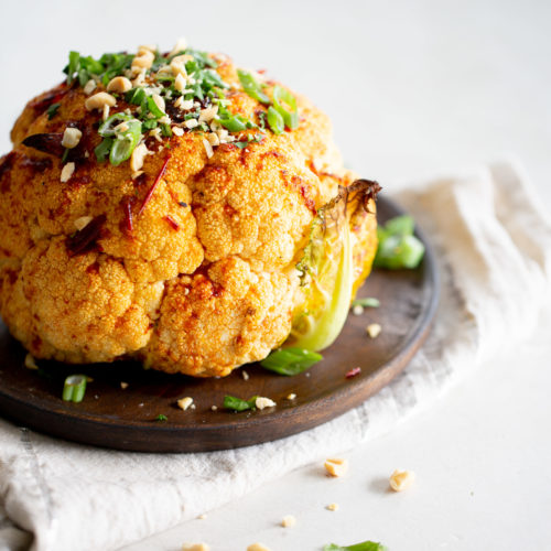 A roasted cauliflower sits on a rustic wooden plate, seasoned with spices and topped with green herbs, chopped nuts, and slices of green onions. A light-colored cloth is underneath the plate, with scattered herbs and nuts surrounding the roasted cauliflower on the white surface.