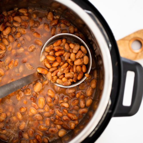 A close-up of an open Instant Pot filled with cooked pinto beans in liquid, perfect for making instant pot refried beans. A metal ladle scoops some beans; the background features part of a wooden cutting board and white countertop.