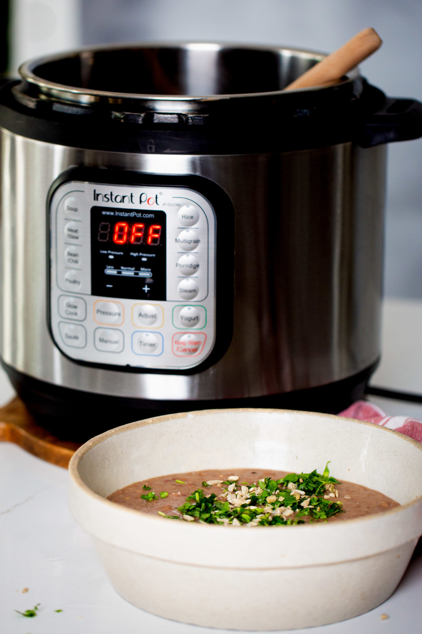 Un tazón de frijoles refritos decorado con hierbas y semillas sobre una encimera blanca frente a una Instant Pot. Una cuchara de madera reposa dentro de la olla, que muestra 'off' en su temporizador digital.