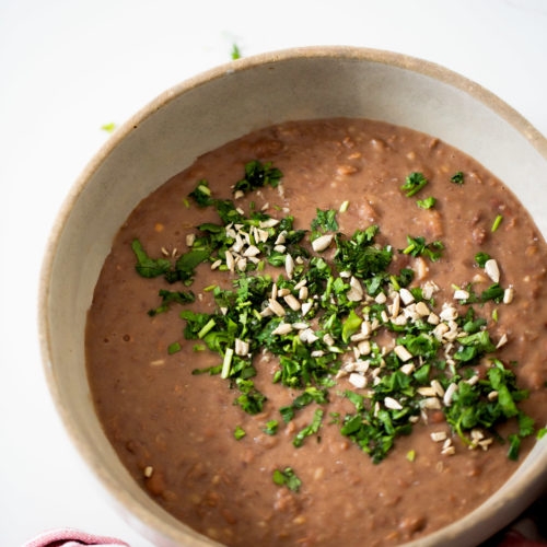A beige bowl filled with creamy #InstantPotBeans refried beans, topped with chopped fresh cilantro and sunflower seeds. The bowl sits on a white surface, partially wrapped in a red and white striped cloth, with scattered herbs nearby.