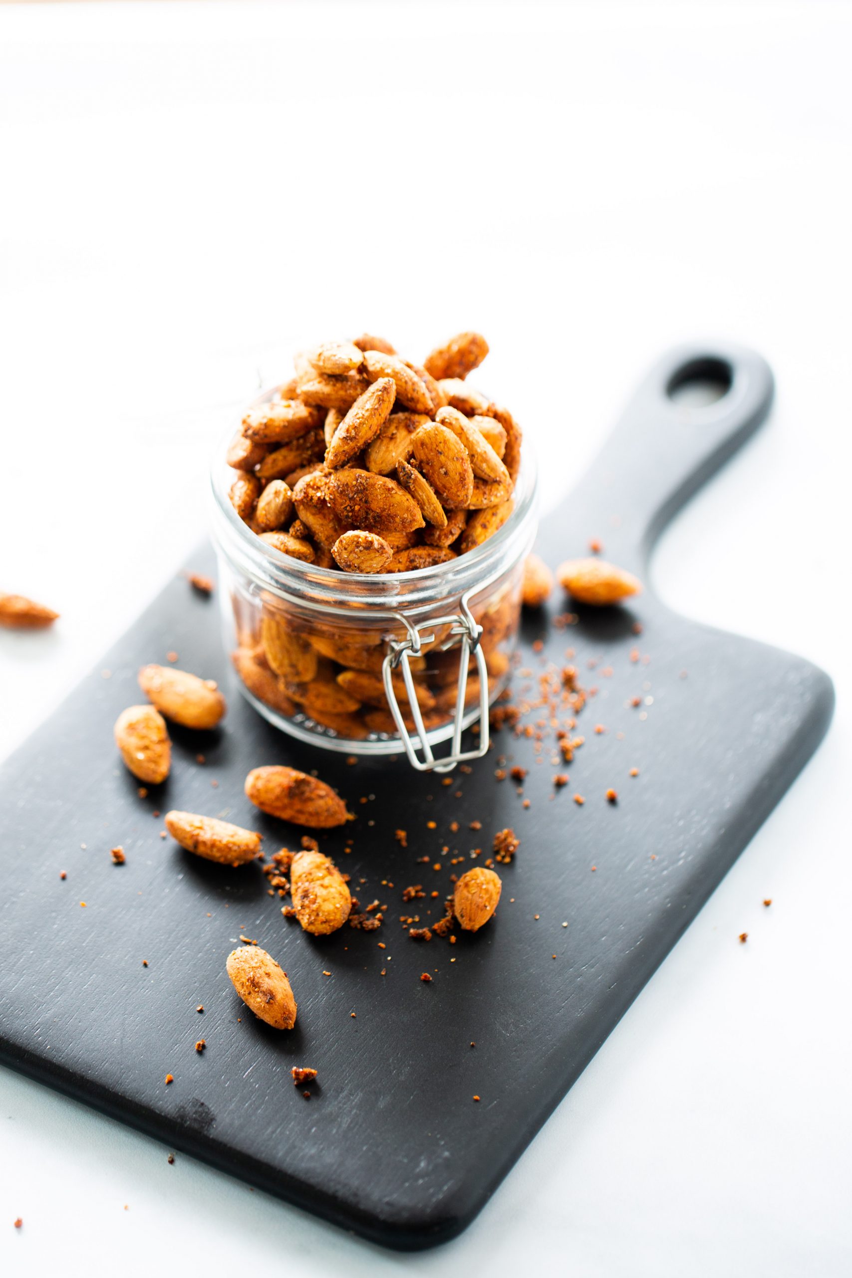Un pequeño frasco de vidrio lleno de almendras veganas tostadas y picantes reposa abierto sobre una tabla de cortar negra. Hay algunas almendras y migas esparcidas por el frasco, mientras que el fondo brillante y ligeramente difuminado destaca estos sabrosos frutos secos.