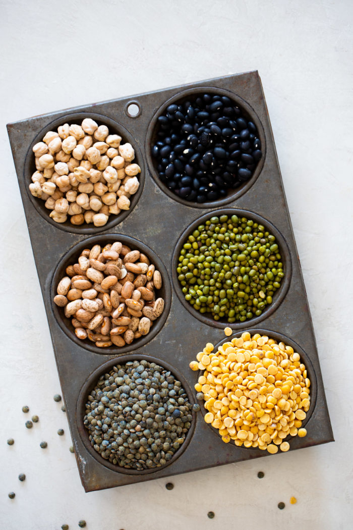 A metal muffin tin with six cups, each filled with a different type of dried legume&mdash;great for anyone ready to cook lentils, beans, or other legumes. Some mung beans are scattered nearby on the light surface.